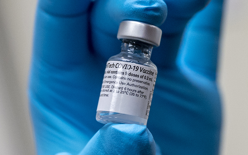 A blue-gloved hand holds a vial of a coronavirus vaccine.