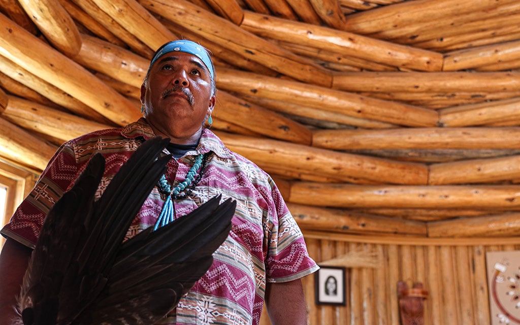 Wayne Wilson stands in a hogan at the Native American Baha’i Institute in Houck, Arizona, on Sept. 1, 2022. He is holding eagle feathers that he uses in traditional healing ceremonies. (Photo by Laura Bargfeld/Cronkite News)