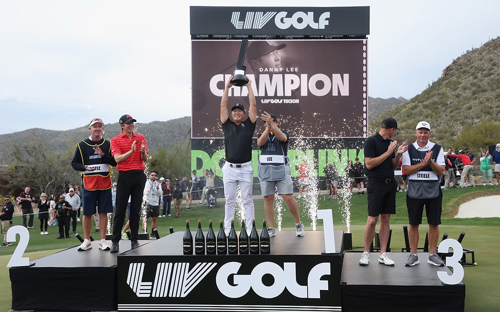 Danny Lee holds the LIV Tucson trophy above his head while standing on the LIV Golf champion podium at a golf course.
