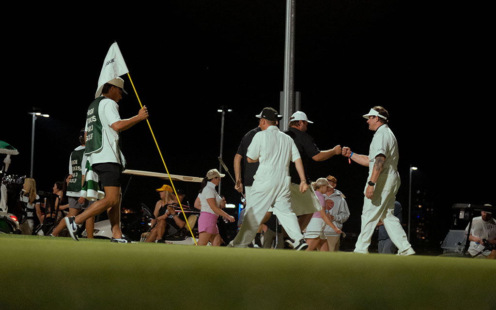 Participants of the inaugural Grass Clippings Open walking across the putting green, one holding a flag indicating the "Grass League," in casual attire under artificial lights.