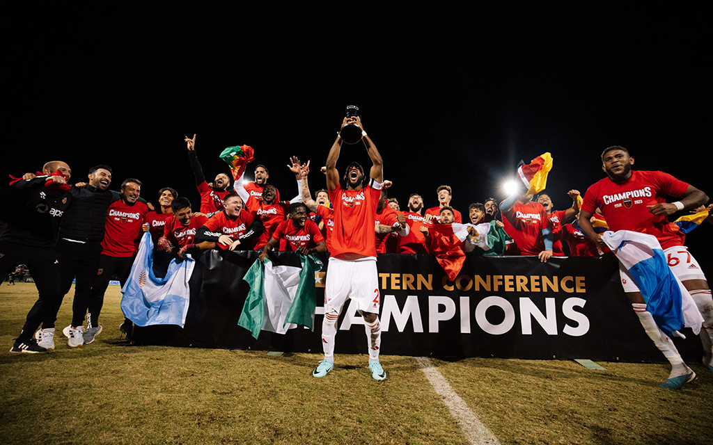 Phoenix Rising FC soccer team celebrating with a trophy in front of a "Champions" banner at night.