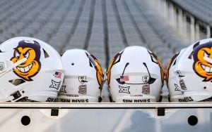 A row of five white football helmets with Sun Devils logo on a metal holder, with one helmet-mounted communication device that allows players to receive and relay play calls directly from coaches on the sideline. Set against a background of empty gray stadium seats.