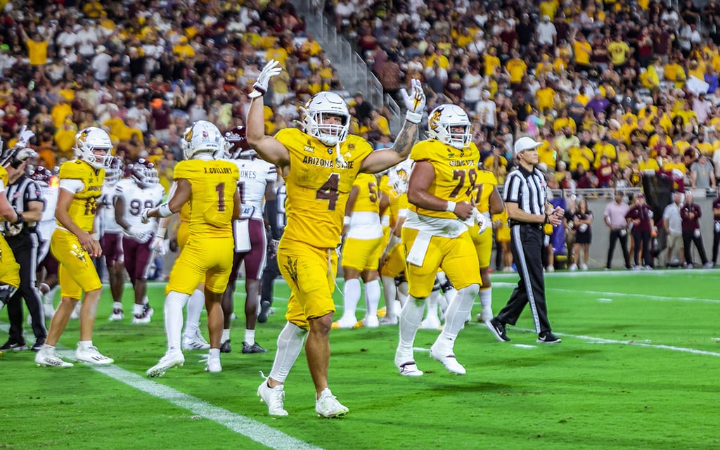 Arizona State football players in yellow uniforms celebrate on the field during a game, with a full stadium of spectators in the background.