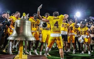 Arizona State in yellow jerseys celebrating near a large silver bell under bright stadium lights.