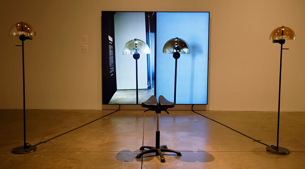 Art installation with three amber dome lamps on black stands, a large screen displaying similar lamps, and a black saddle-style stool in the foreground.