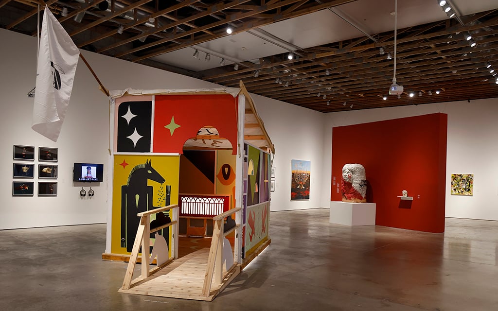 Interior of a modern art gallery with diverse artworks, including a colorful house-like installation and multiple framed photographs. The installation is made of panels have geometric designs, including a black horse-like figure on a yellow background. The house has an archway entrance and a small wooden ramp in front. To the left of the installation, a white flag is hanging from a wooden beam.