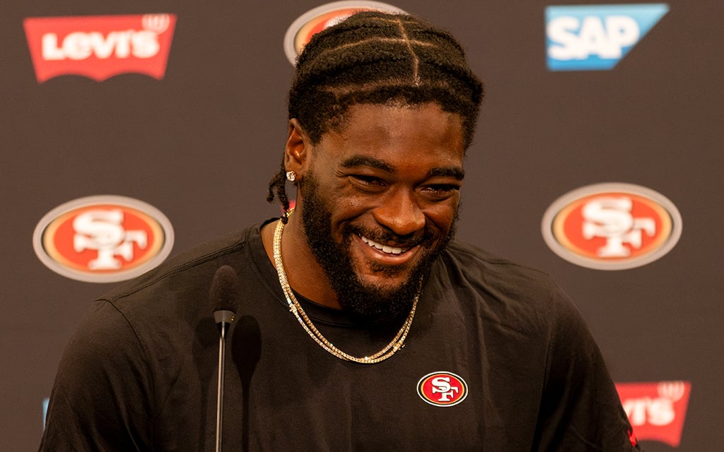 Brandon Aiyuk is wearing a black t-shirt and a gold chain stands at a podium with San Francisco 49ers, LEVI'S, and SAP logos in the background during a press conference.