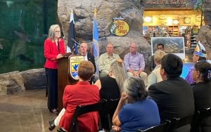 Katie Hobbs in a red blazer speaks at a podium with the "Fish & Wildlife Service" seal at an indoor event beside an aquarium, with an audience and seated panelists, including a uniformed man; the "Bass Pro Shops" sign is visible in the background.