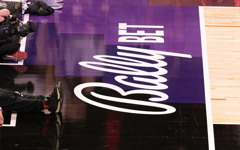 A basketball court's glossy wooden floor with a bold "Bally BET" advertisement in white on a purple background, including a glimpse of courtside equipment and people.