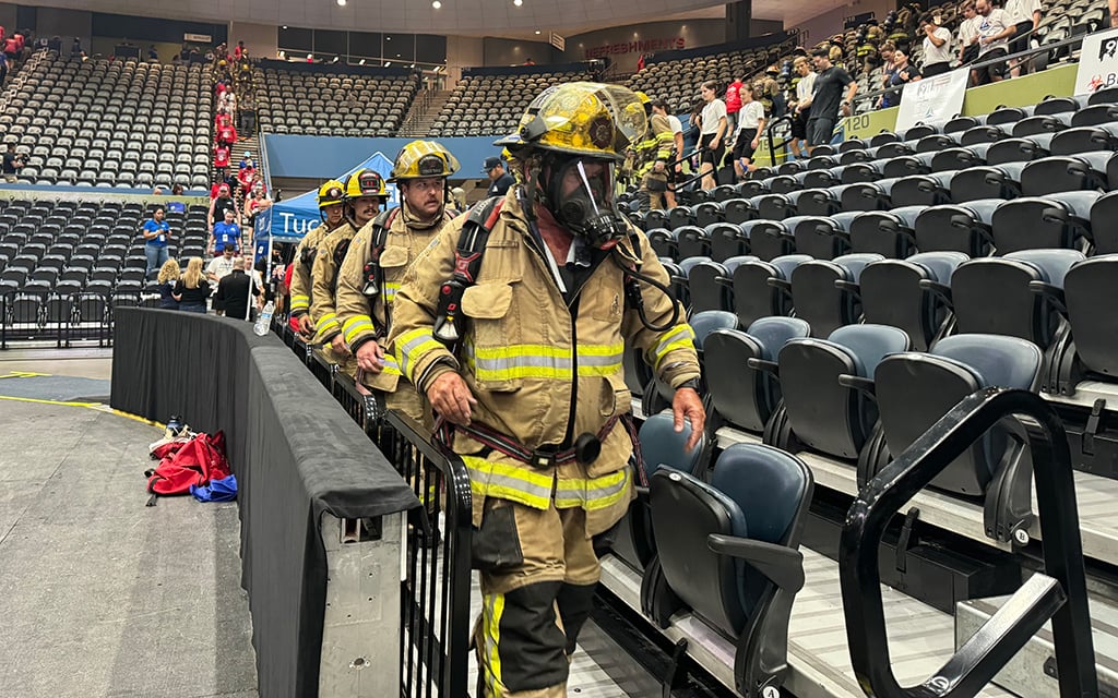 Members of the Tucson Fire Department firefighters in protective gear walk single file down a stairway in a large indoor arena with empty and occupied seats.