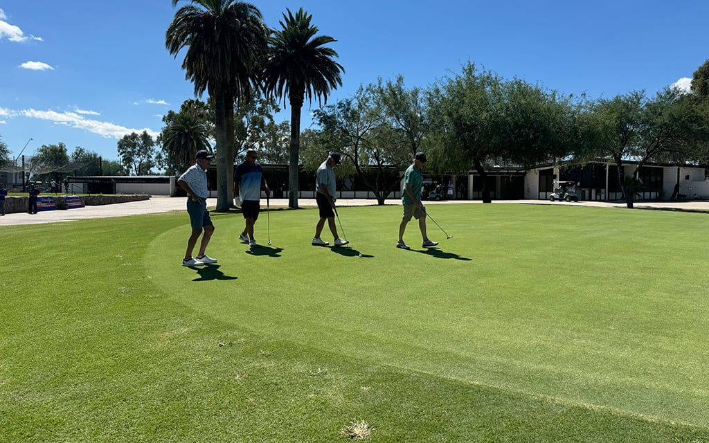 Four people on a green golf course holding putters with palms and buildings in the background under a clear blue sky.