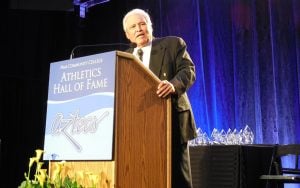 Rich Alday, who died in 2021, speaking at a podium with a sign reading "Pima Community College Athletics Hall of Fame Aztecs," with yellow flowers and crystal awards nearby.