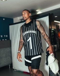 Isaiah Thomas in black and white striped sports jersey marked "SLOWGRIND" and shorts, standing indoors holding a white fabric item.