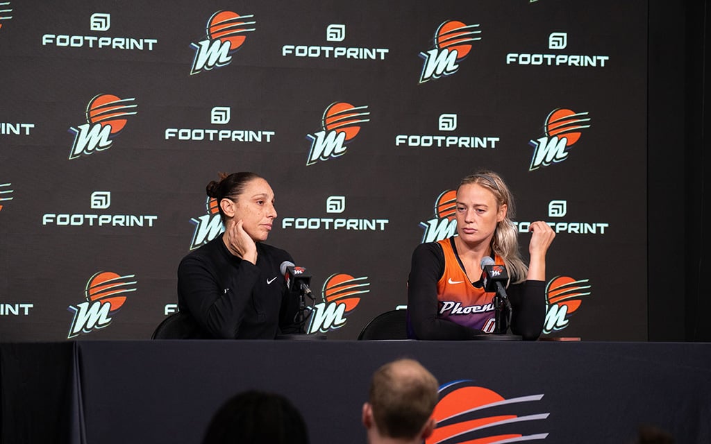 Diana Taurasi and Sophie Cunningham seated at a press conference table with microphones, in front of a backdrop featuring repeated logos and the word "Footprint."