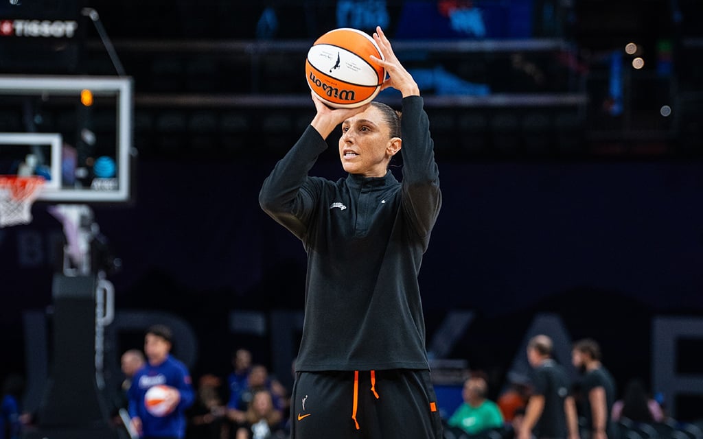 Diana Taurasi in black and orange athletic attire preparing to shoot a basketball on an indoor court, with a blurred backdrop of basketball hoops and spectators.