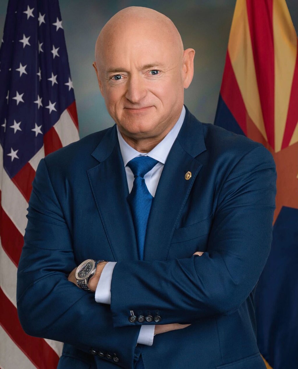 Official portrait of Arizona Senator Mark Kelly in a blue suit with arms crossed in front of a U.S. flag and an Arizona state flag.