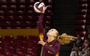 Mary Shroll in a maroon and gold jersey serving the ball in a stadium.