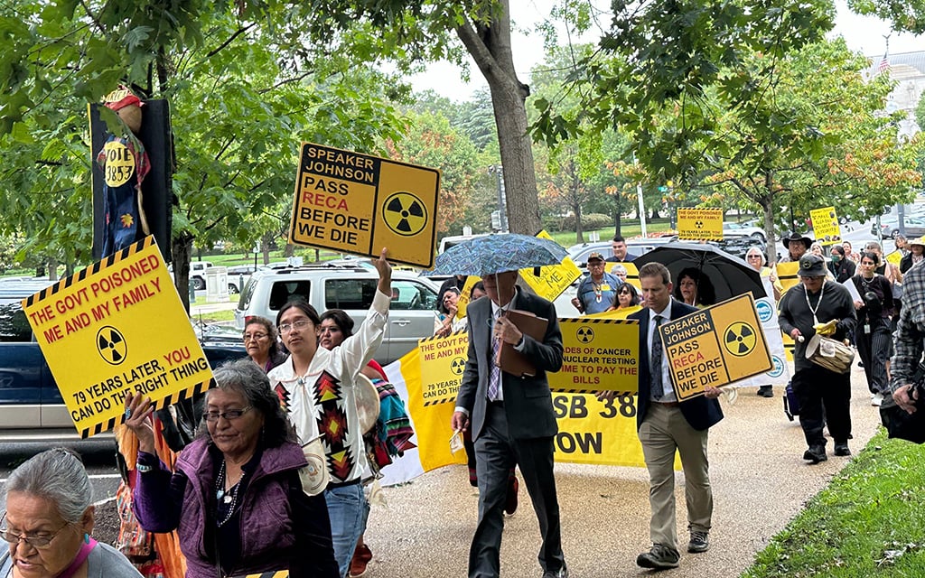 People marching in a protest holding yellow signs with black text and radiation symbols.