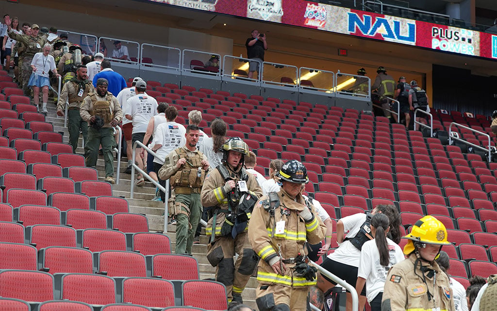 Two firefighters in full gear standing beside a brass bell on a wooden frame in a stadium.