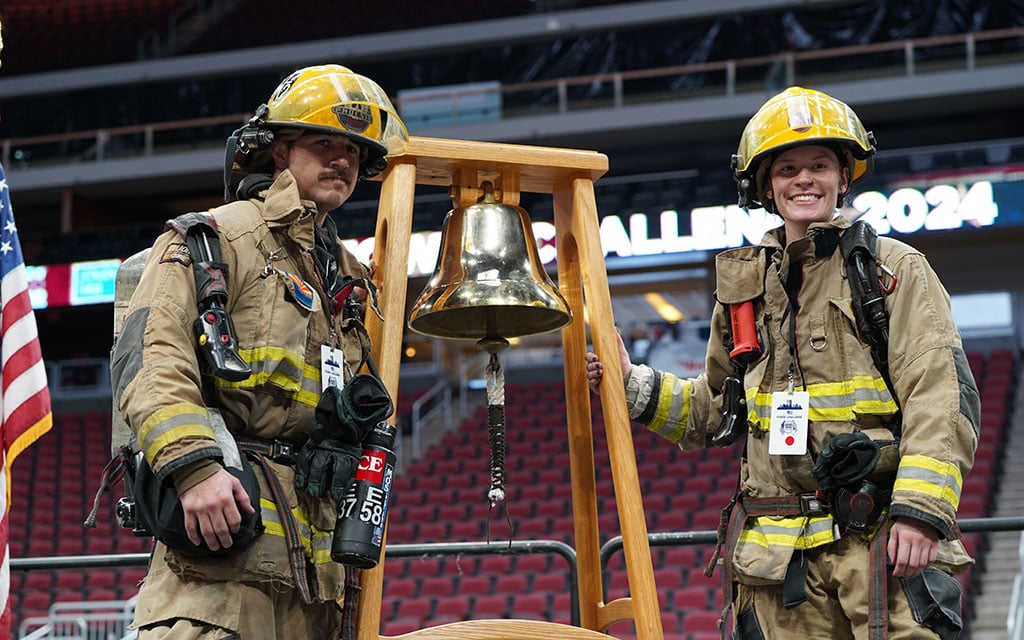 Two firefighters in full gear standing beside a brass bell on a wooden frame in a stadium.