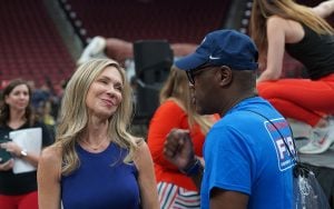 A woman in a blue top smiles at a man in a blue T-shirt and cap in an indoor stadium setting.