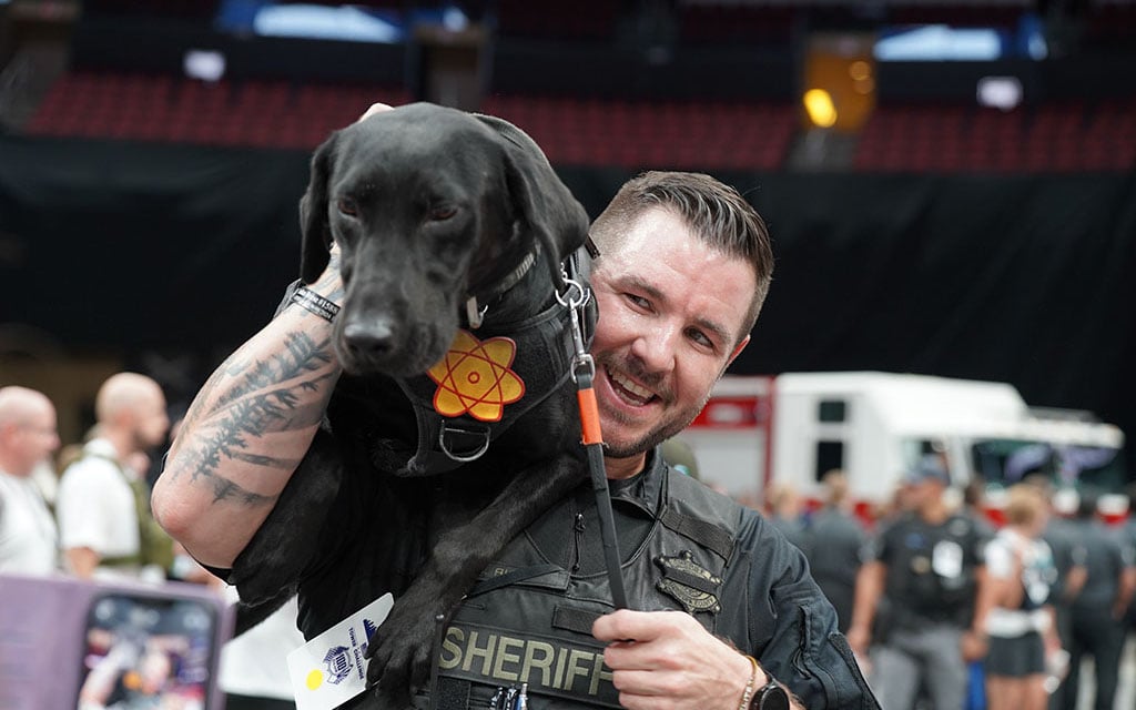 Man in a sheriff's uniform holds a black dog wearing a harness with an atomic symbol.