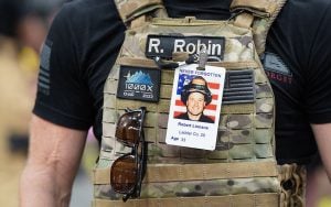 Person wearing camouflage tactical vest with a name tag "R. Robin", patches, sunglasses, and a laminated card displaying a man’s photo with firefighter gear.