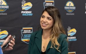 Regina Sirvent in a dark green blazer smiles while being interviewed. Logos for Phoenix Raceway and NASCAR events are visible in the background.