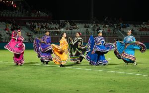 Seven dancers in colorful traditional Mexican dresses perform at Phoenix Rising Stadium, with spectators in the background.