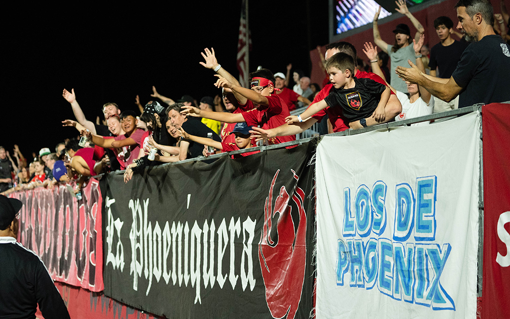 Enthusiastic fans at a Phoenix Rising FC match behind a barrier with banners.