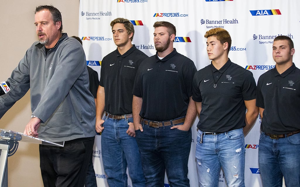 Steve Campbell speaking at a podium with four young men standing in a row next to him, all dressed in black collared shirts and blue jeans, in front of a white background with multiple logos.