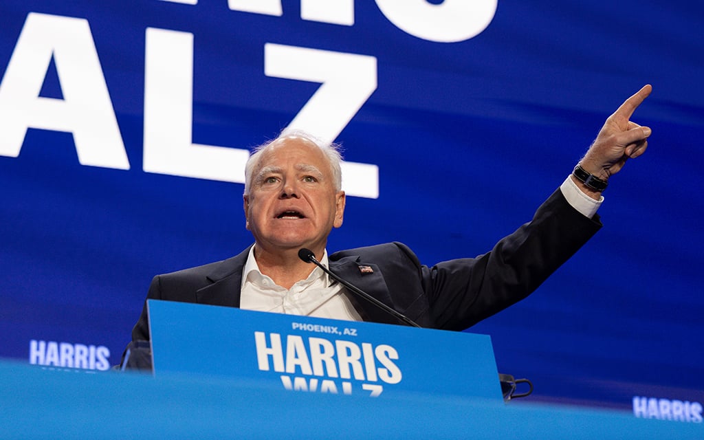 Tim Walz in formal attire speaking at a podium with a blue background and white text.