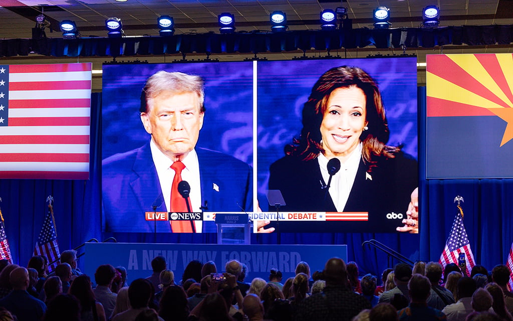 Two large video screens at a public event portray former President Donald Trump on the left and Vice President Kamala Harris on the right, viewed by an audience. American and Arizona flags are displayed in the background.