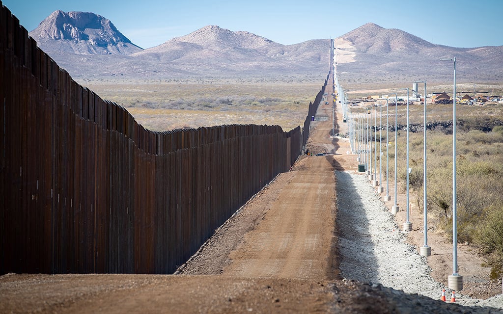 A long metal border fence extends through a desert, accompanied by a dirt road lined with tall poles.