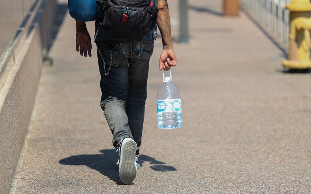 Person walking on a sidewalk carrying a large plastic jug of water, a sleeping bag, and wearing a backpack.