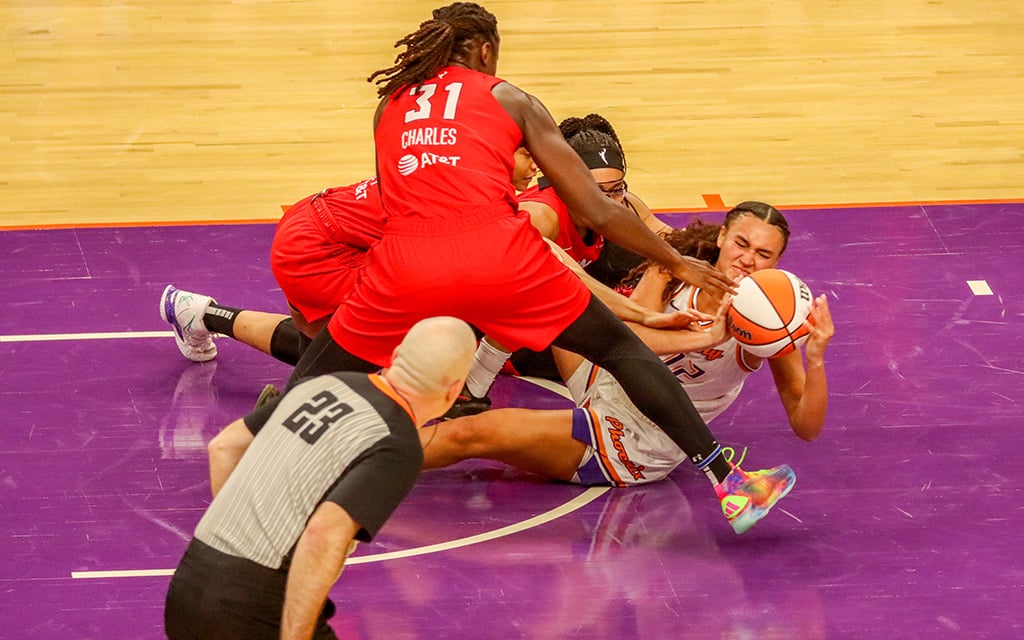 Celeste Taylor tussles for the basketball on the court during a WNBA basketball game, while a referee looks on.