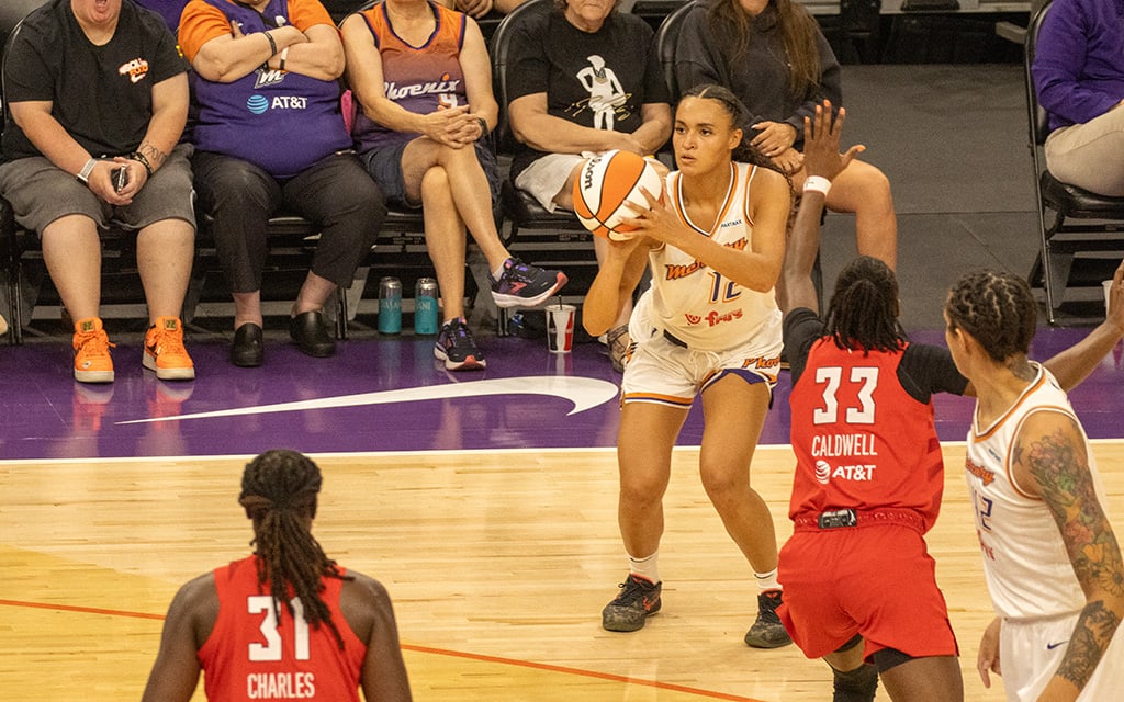 Celeste Taylor shoots the ball, facing an opponent in red uniform during a game, with spectators in the background.