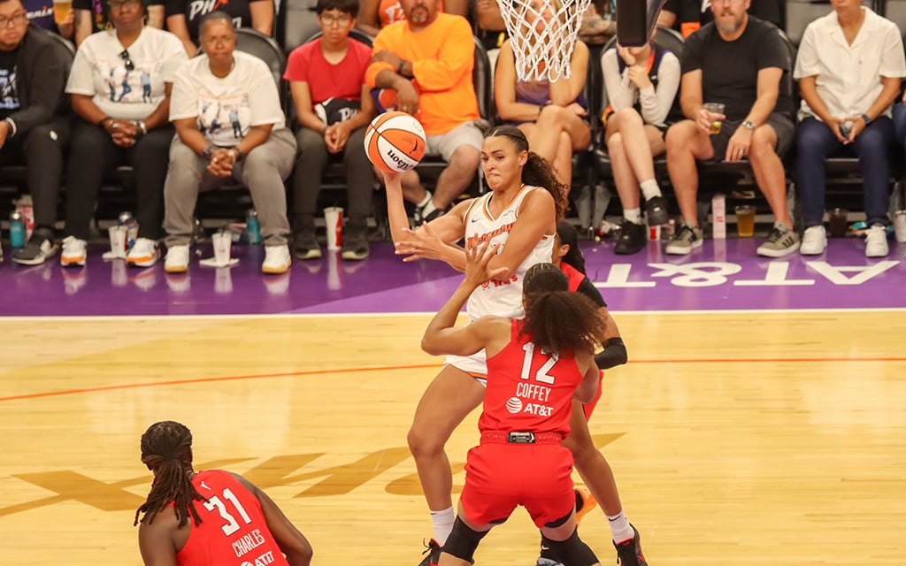 Phoenix Mercury guard Celeste Taylor passes a basketball while being guarded by a player in a red jersey. Spectators in the background intently watch the game.