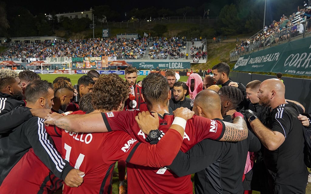 Phoenix Rising FC players and coaches huddled on the field with a stadium full of spectators in the background.