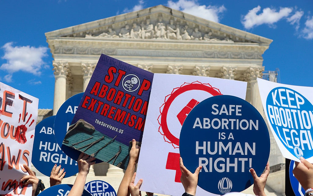 Protesters holding up signs with messages about abortion in front of the U.S. Supreme Court building on a clear day.