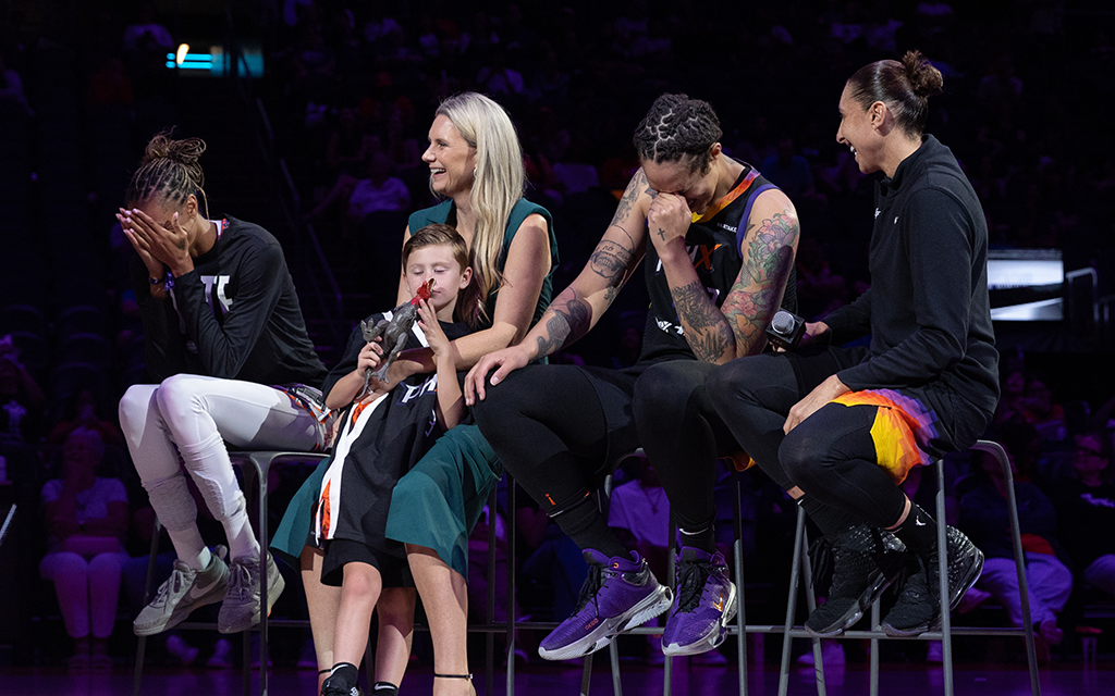 A group of five Phoenix Mercury members seated on stage chairs under focused lighting, with two covering their faces, one smiling, and a child holding a stuffed animal.