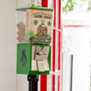 A green vending machine for seed bombs with a coin slot and dispensing lever.