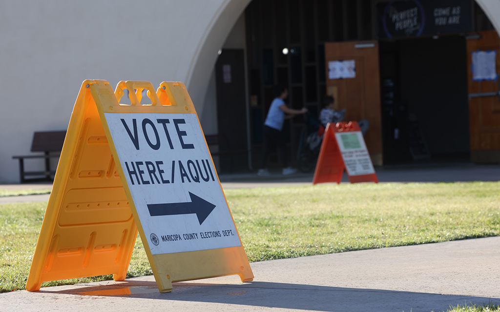 Yellow "Vote Here" sign on a lawn near a building entrance.