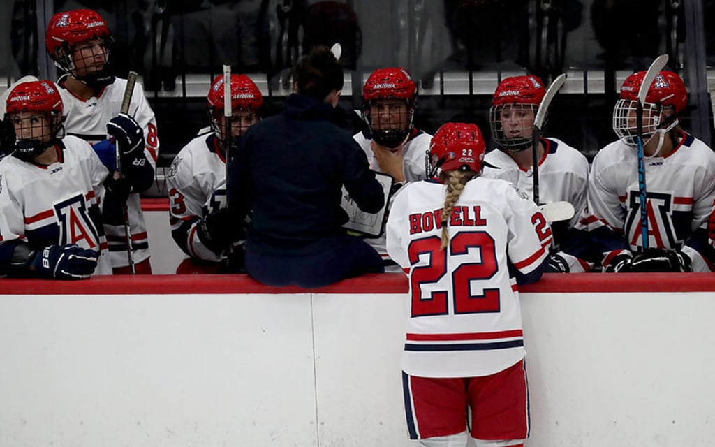 University of Arizona women's ice hockey team on the bench, with coach Caitlin Hogan giving instructions.