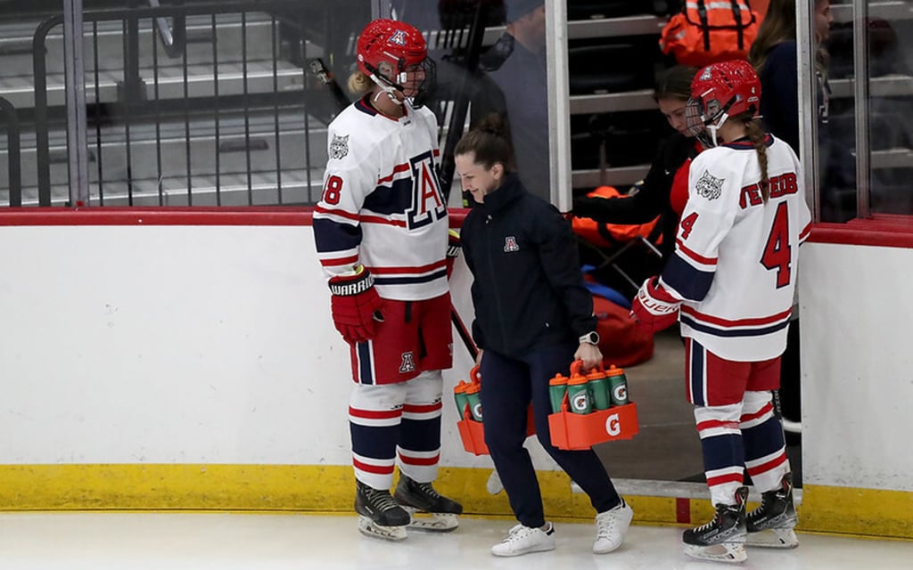 Arizona Wildcats women's ice hockey players in white jerseys stand rink-side as coach Caitlin Hogan carries a tray of Gatorade bottles.