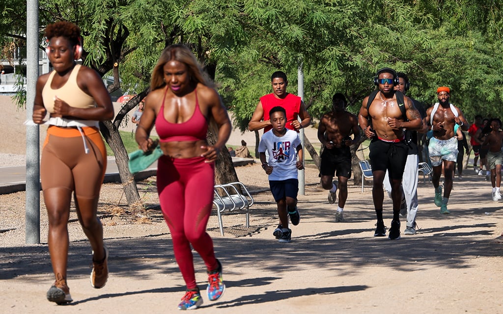 Group of people in the BLK Arizona Run Club running on a dirt path at Tempe Town Lake with trees and benches on a sunny day.