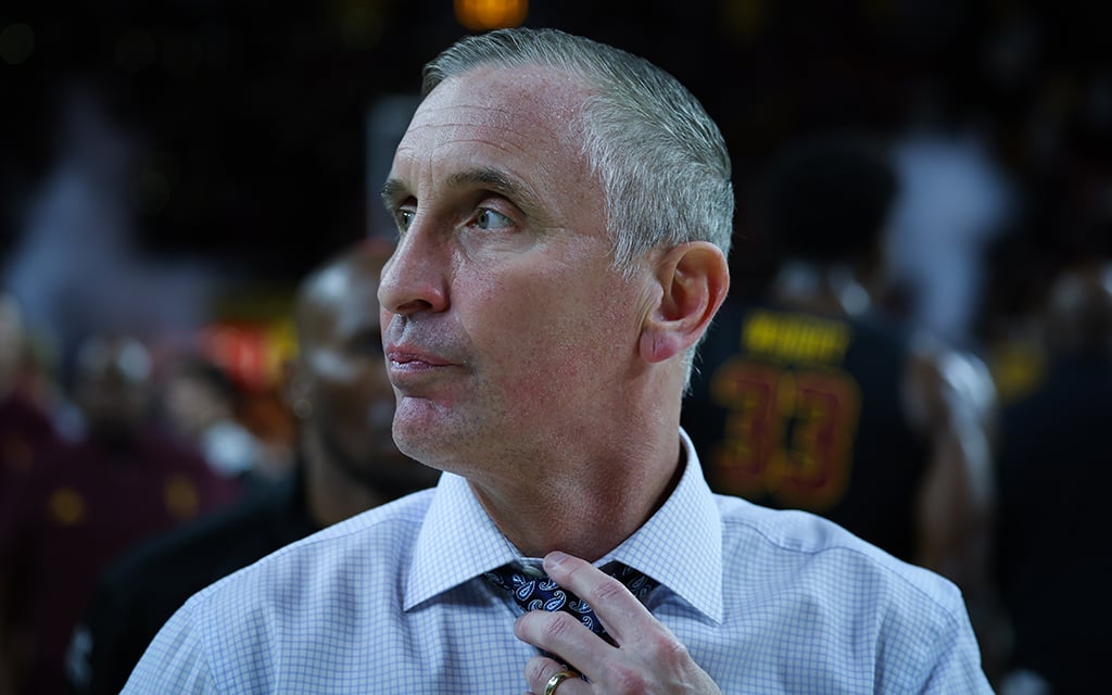 Arizona State basketball coach Bobby Hurley in a suit adjusting their tie in a dimly lit sports setting.