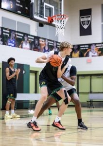 Three basketball players during practice, with one holding a ball and being closely guarded.