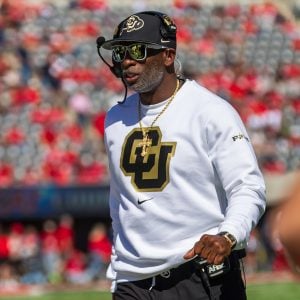 Man in a white sweatshirt with a "CU" logo, wearing a headset and mirrored sunglasses, walks on a sports field.