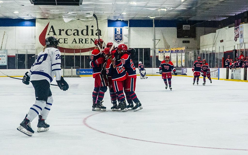 Arizona Wildcats women's ice hockey players in red and blue uniforms celebrate in the rink, with another player in white and blue skating towards them.